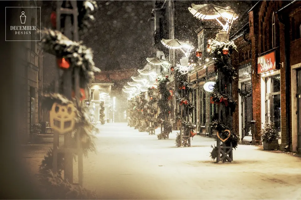 A row of wooden poles in a snowy street, each pole wrapped in a garland of needles, red balls and wooden heart shapes, bright fan-shaped light sculptures above