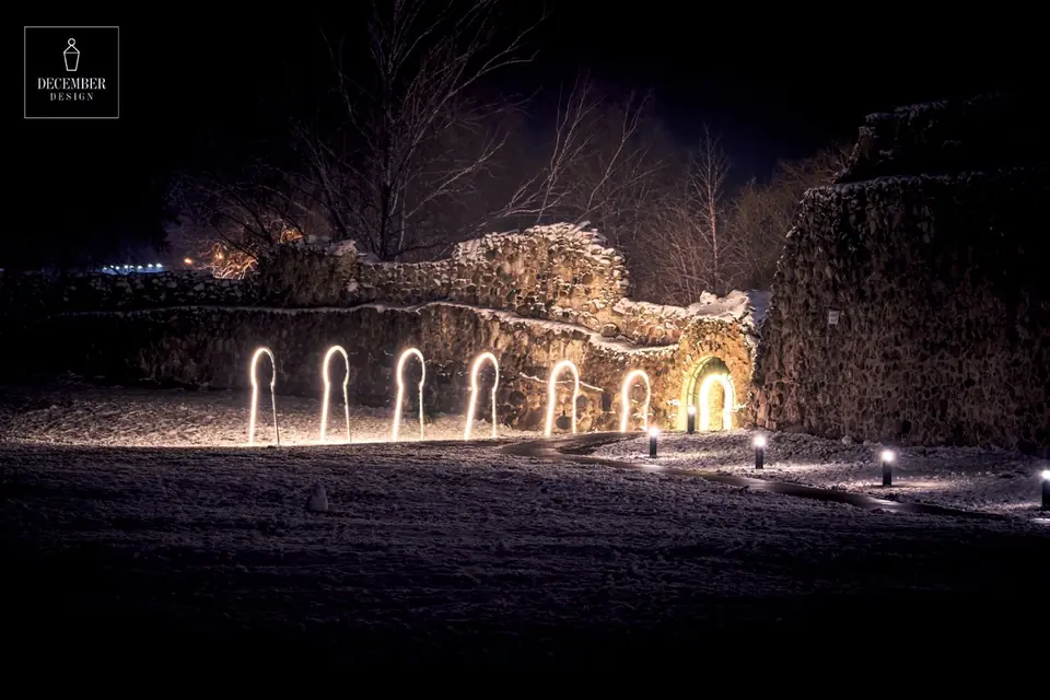 A row of glowing arches of light along the snow-covered walls of Dobele Castle leading to an illuminated door