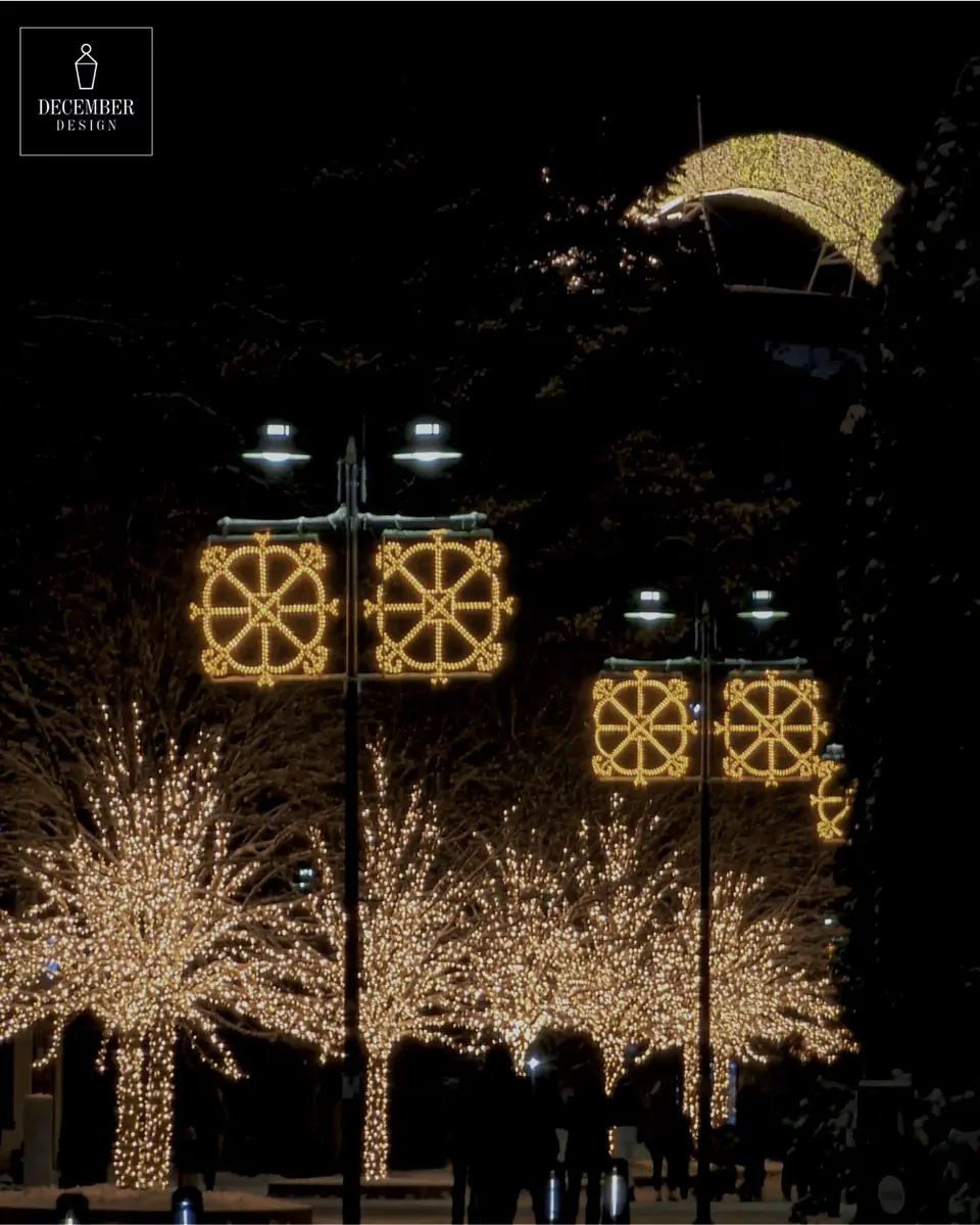 Street lamps with square star decorations and trees covered in warm white light on a snowy street at night