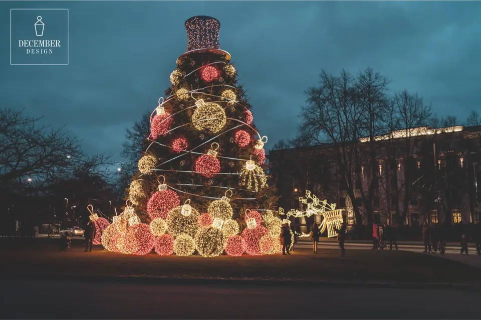 Christmas tree with spiral strings of lights, red and white balls and an illuminated cylinder top in a park near a historic building