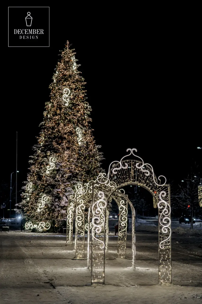 Three illuminated arches with a scroll pattern form a short tunnel, with a tall Christmas tree in the background