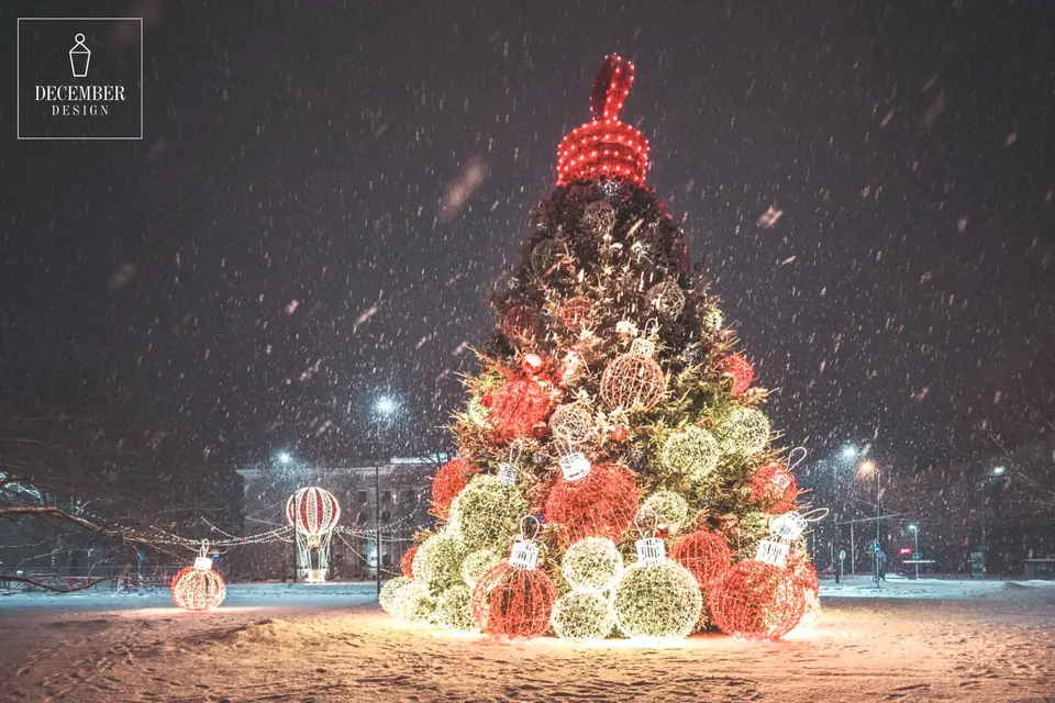 A 12-metre-high Liepāja fir tree, decorated with red and warm white light balls during snowfall, an illuminated hot air balloon can be seen next to it