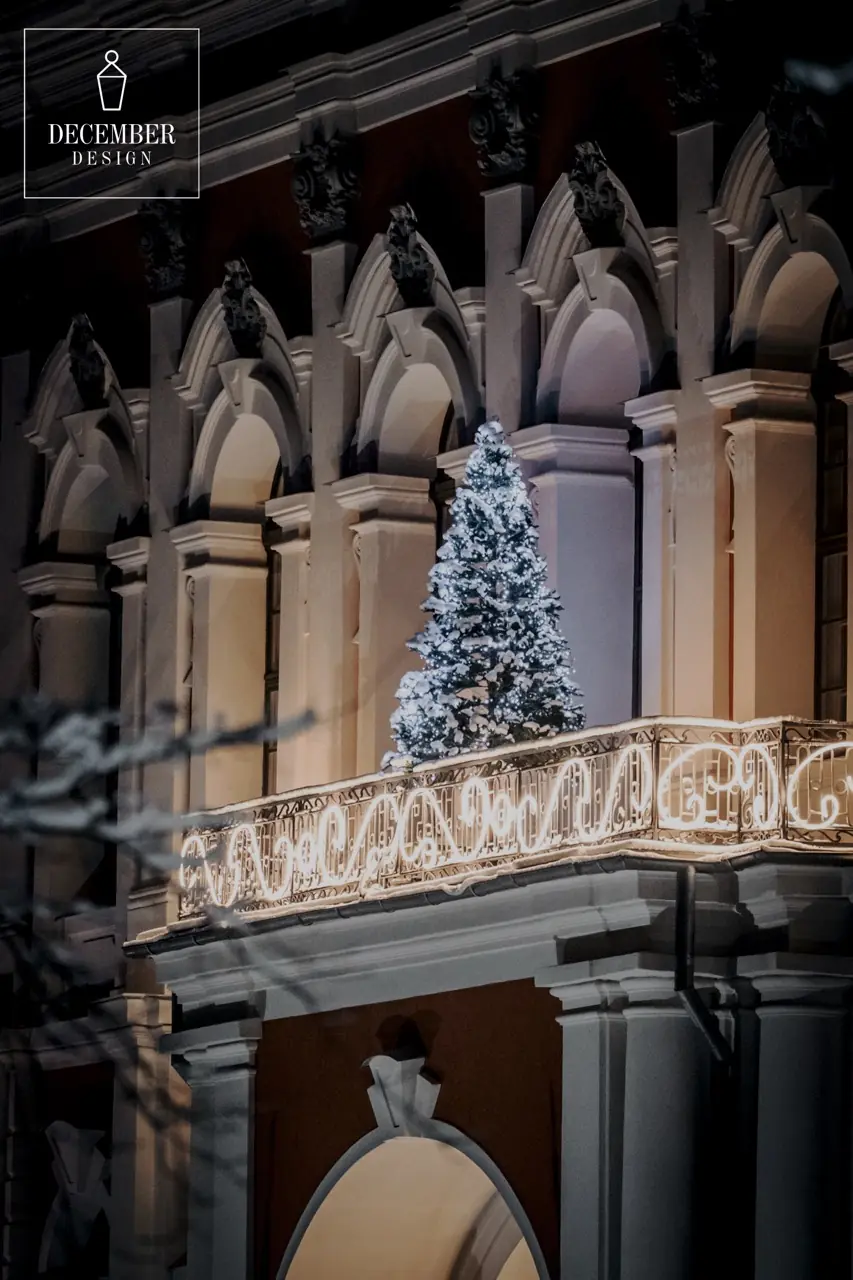 Snow-covered, white-lit Christmas tree on the balcony of a classic building with light decorations