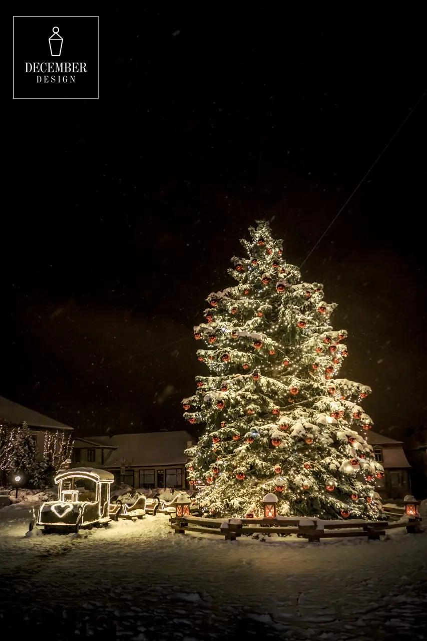 Night view of a snowy town square with a large Christmas tree decorated with warm lights and red balls and a wooden train light installation