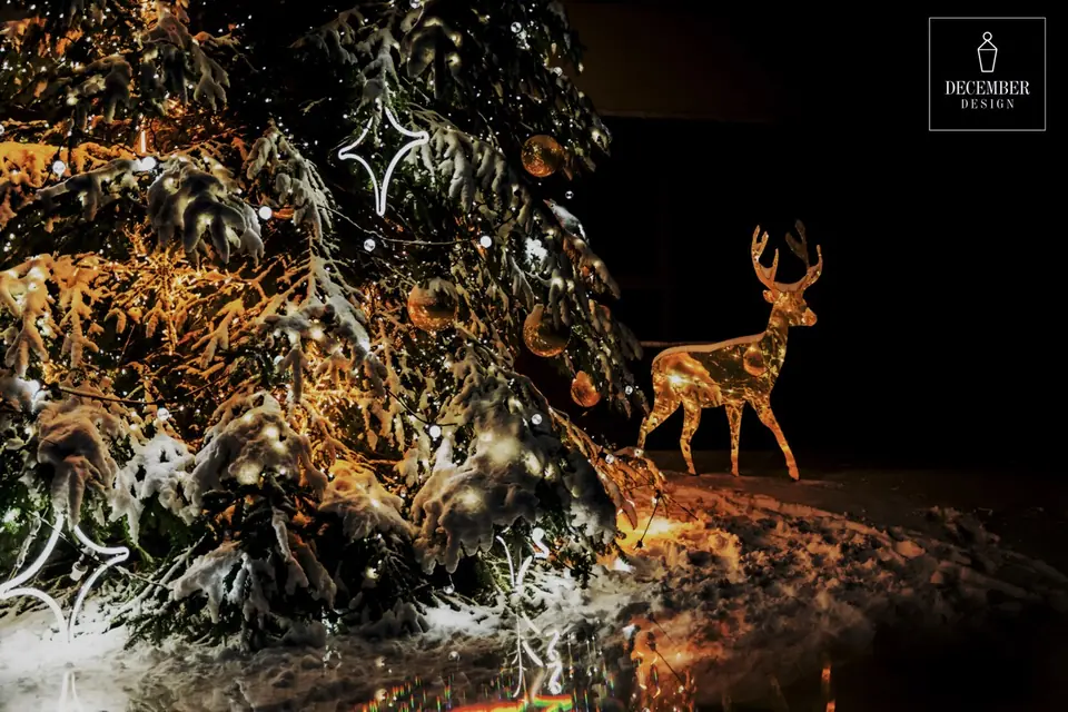 A snowy, illuminated Christmas tree with golden balls and a glittering deer sculpture next to it
