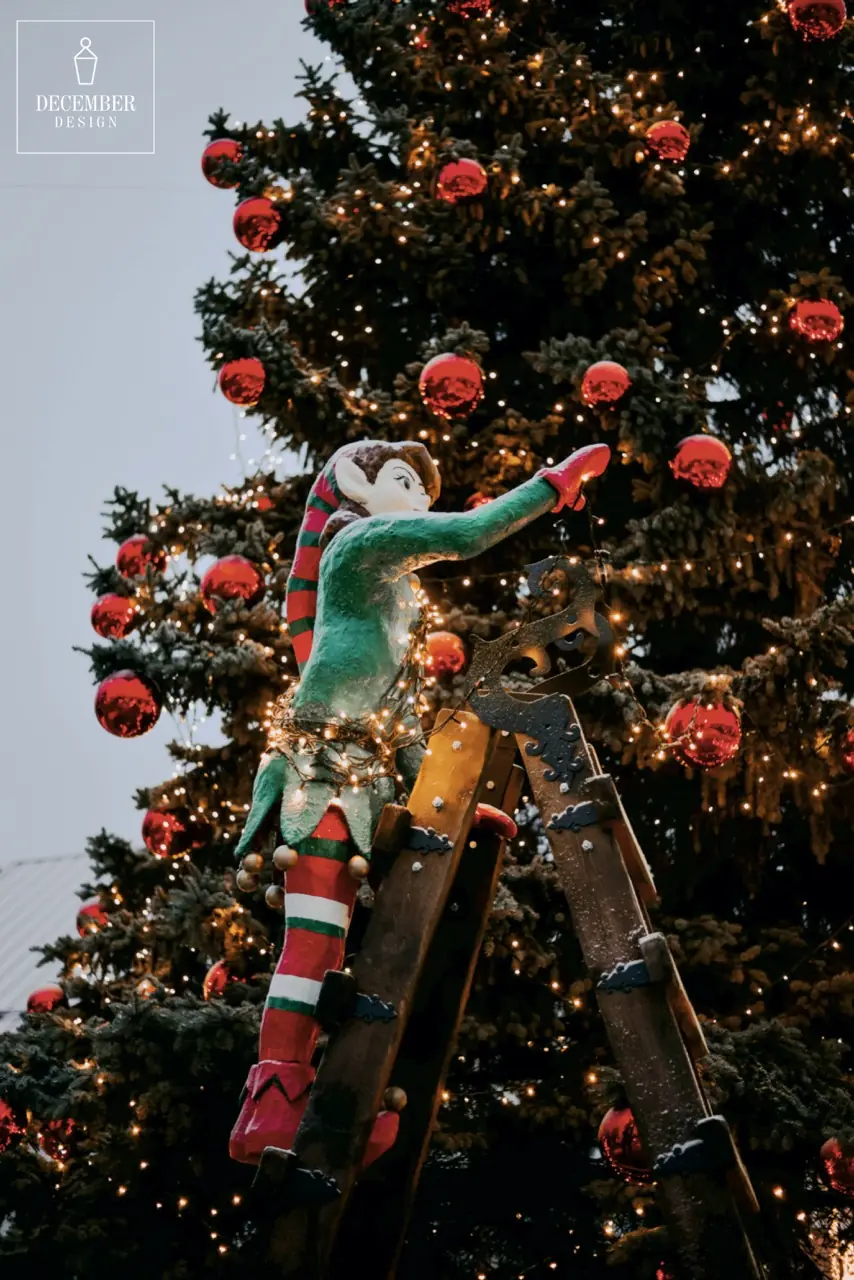 A gnome on a wooden staircase decorates a large Christmas tree with warm lights and red balls in daylight