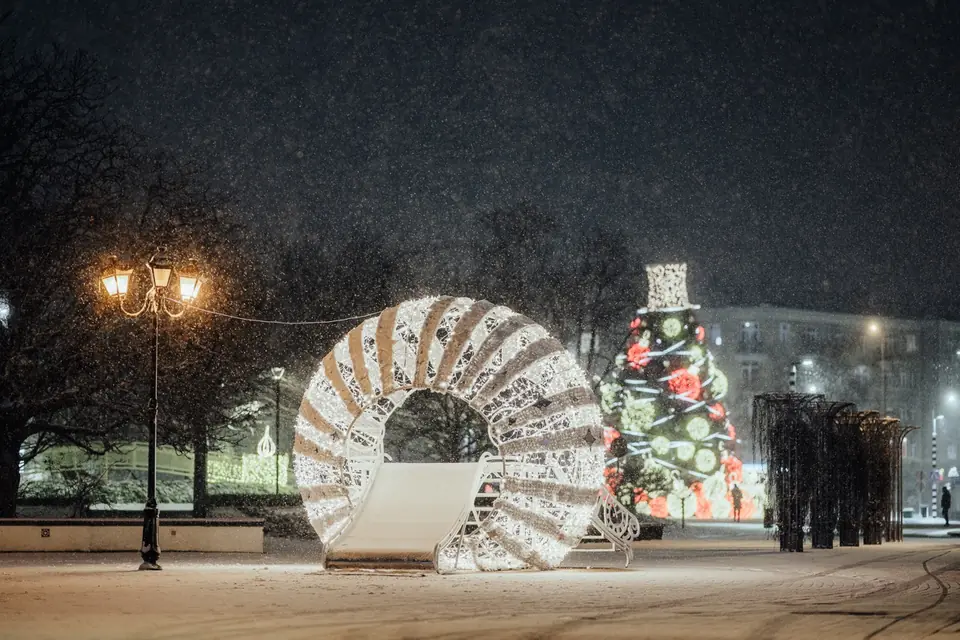 A large circular light tunnel with a snow-covered skating rink inside, a multi-tiered Christmas tree with red and green lights in the background