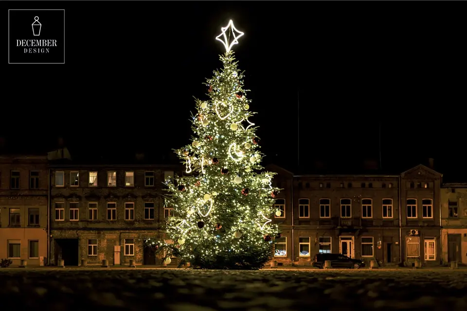 A large Christmas tree with warm white lights, heart and moonlight figures and a large white star on top, placed in a cobbled square near old buildings