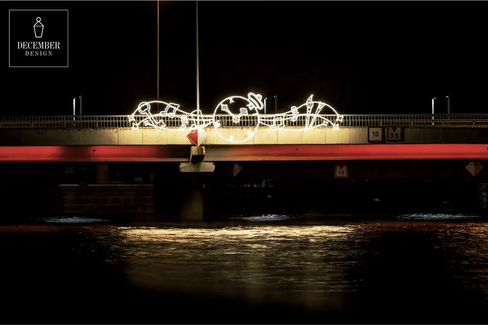 A bridge over a dark body of water, the edges of which are dressed in white light outlines with clock, bird and scroll motifs, a red light bar along the bridge